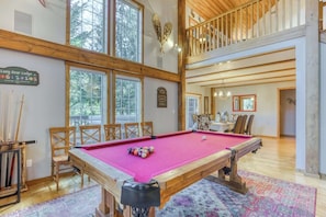 A pool table in a spacious room with wooden beams, a dining area, and a view of trees through large windows. - Lazy Bear Lodge: Hot Tub, Game Room & Dogs OK (Mount Hood Village)