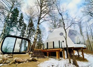 Eine geodätische Kuppel in bewaldeter Landschaft mit einer Holzterrasse und Treppen, die zu ihr führen.