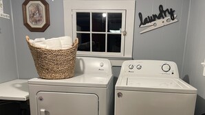 A laundry area with two white front-loading washers and dryers, a wicker basket on the left washer, a window above the dryers, and a 'Laundry' sign on the wall.