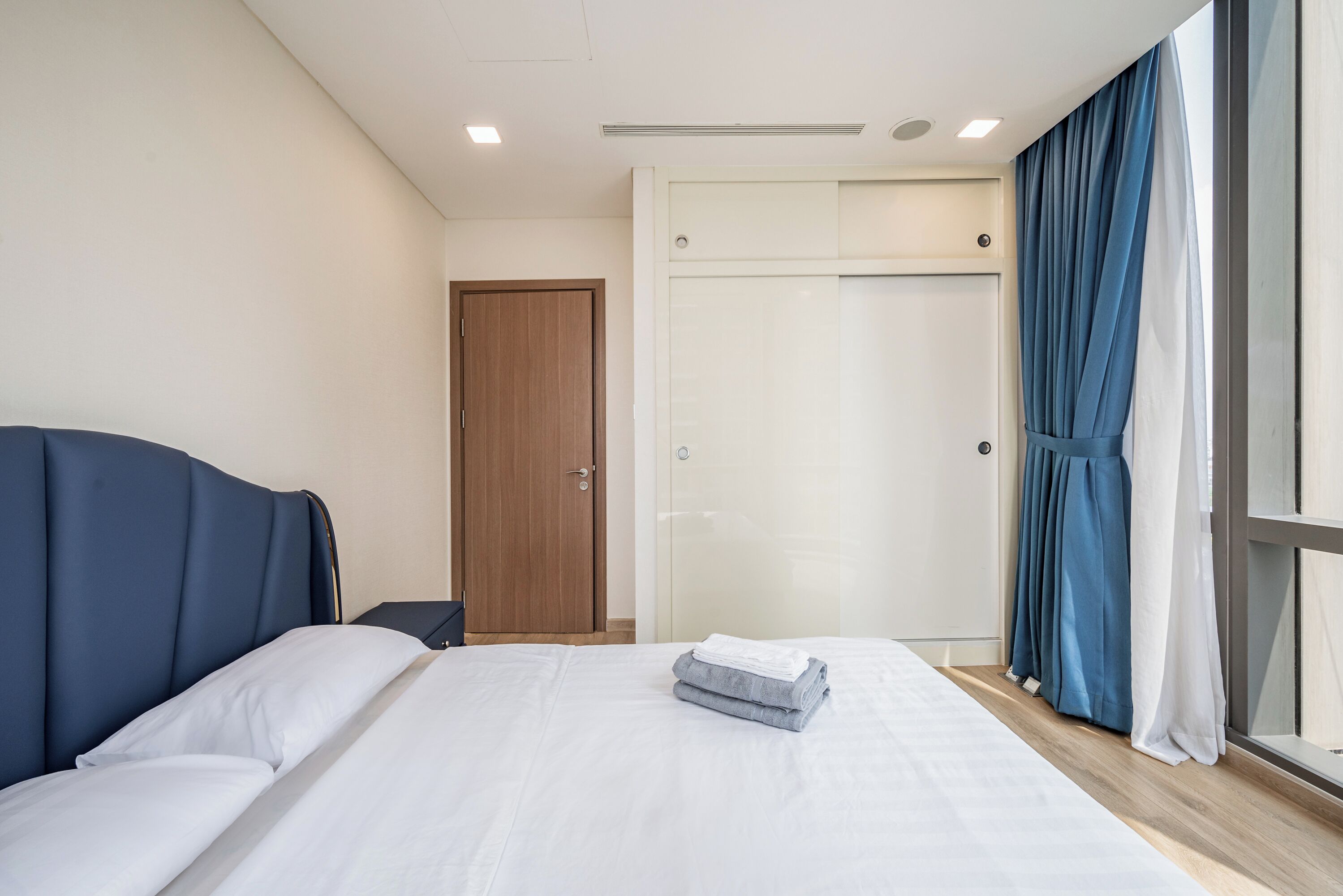 Un lit bien fait, avec du linge de lit blanc et une serviette pliée, une porte en bois et une fenêtre ornée de rideaux bleus.