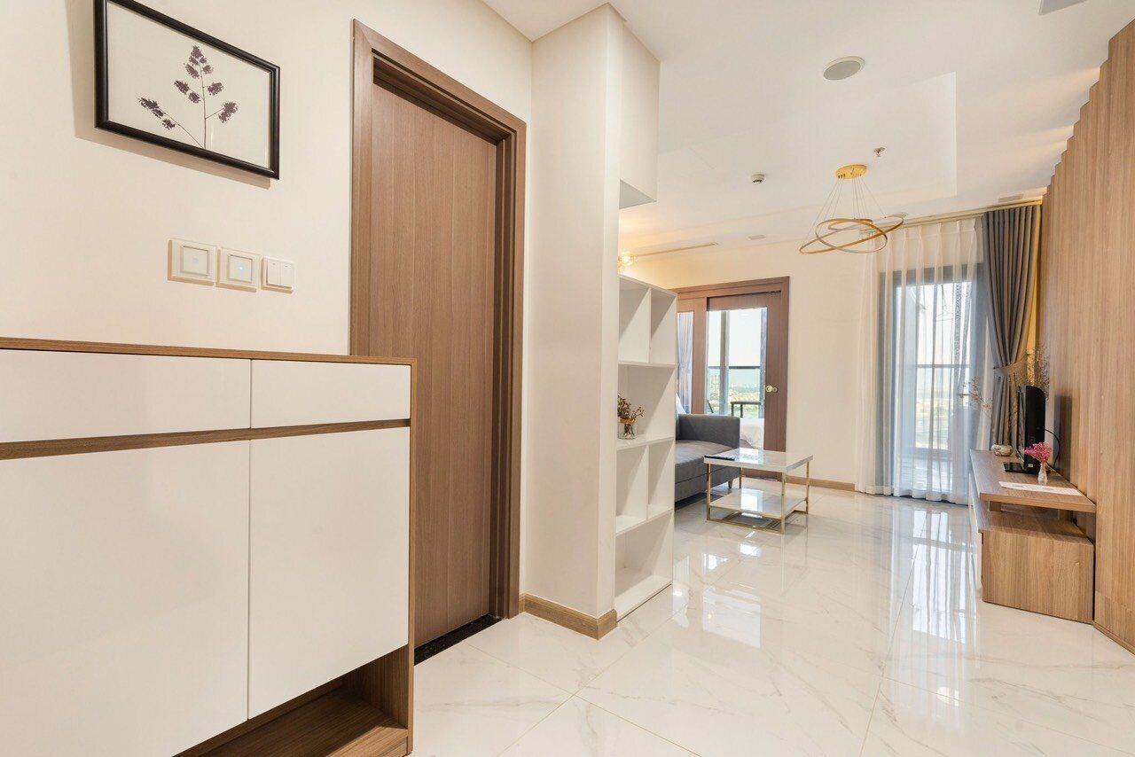 Un interior moderno con una puerta de madera, armarios blancos, una mesa de centro de vidrio y vistas a una sala de estar con un sofá y cortinas.