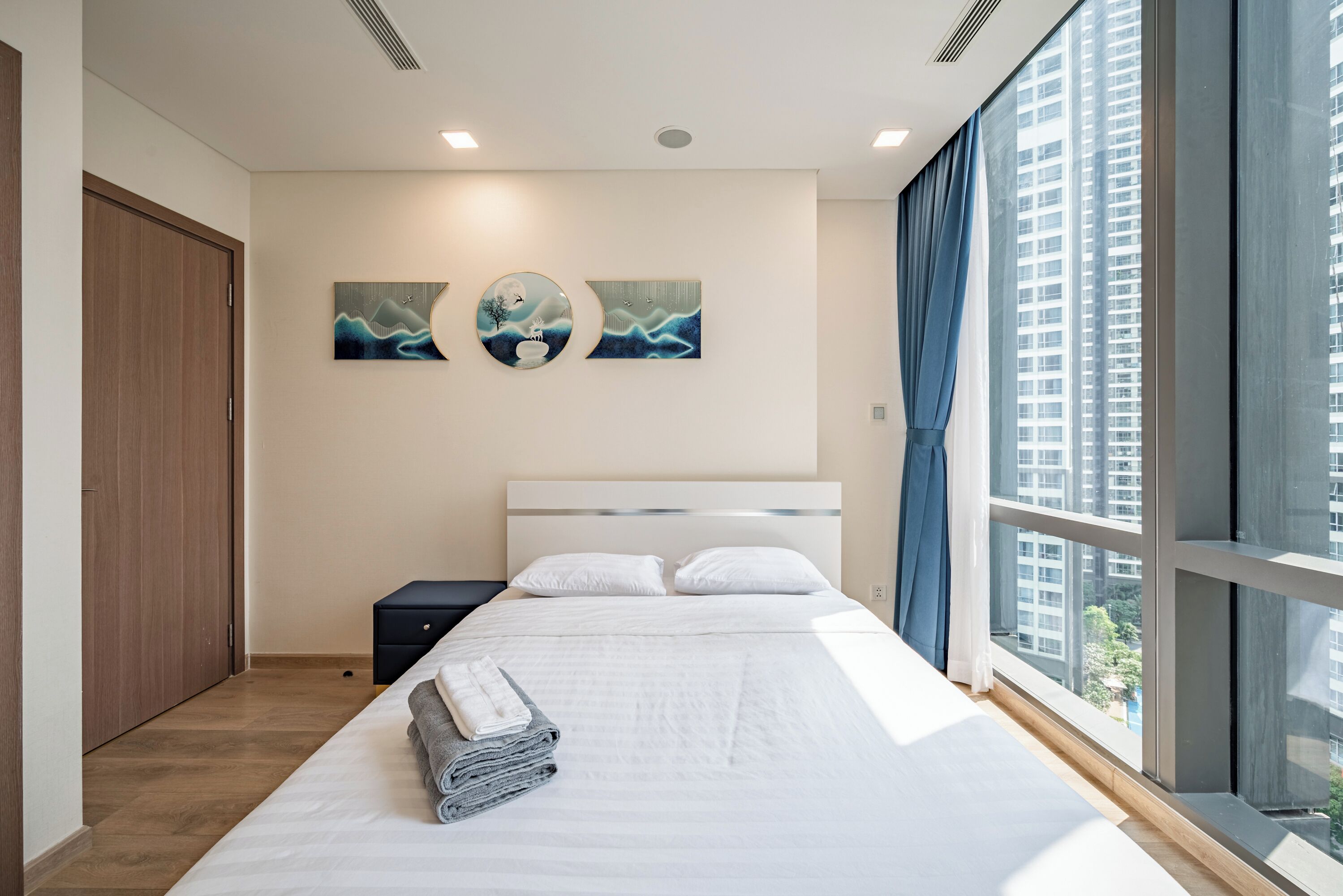 Une chambre d’hôtel moderne, dotée d’un grand lit, d’une table de chevet, d’une armoire et offrant une vue sur la ville grâce à de grandes fenêtres.