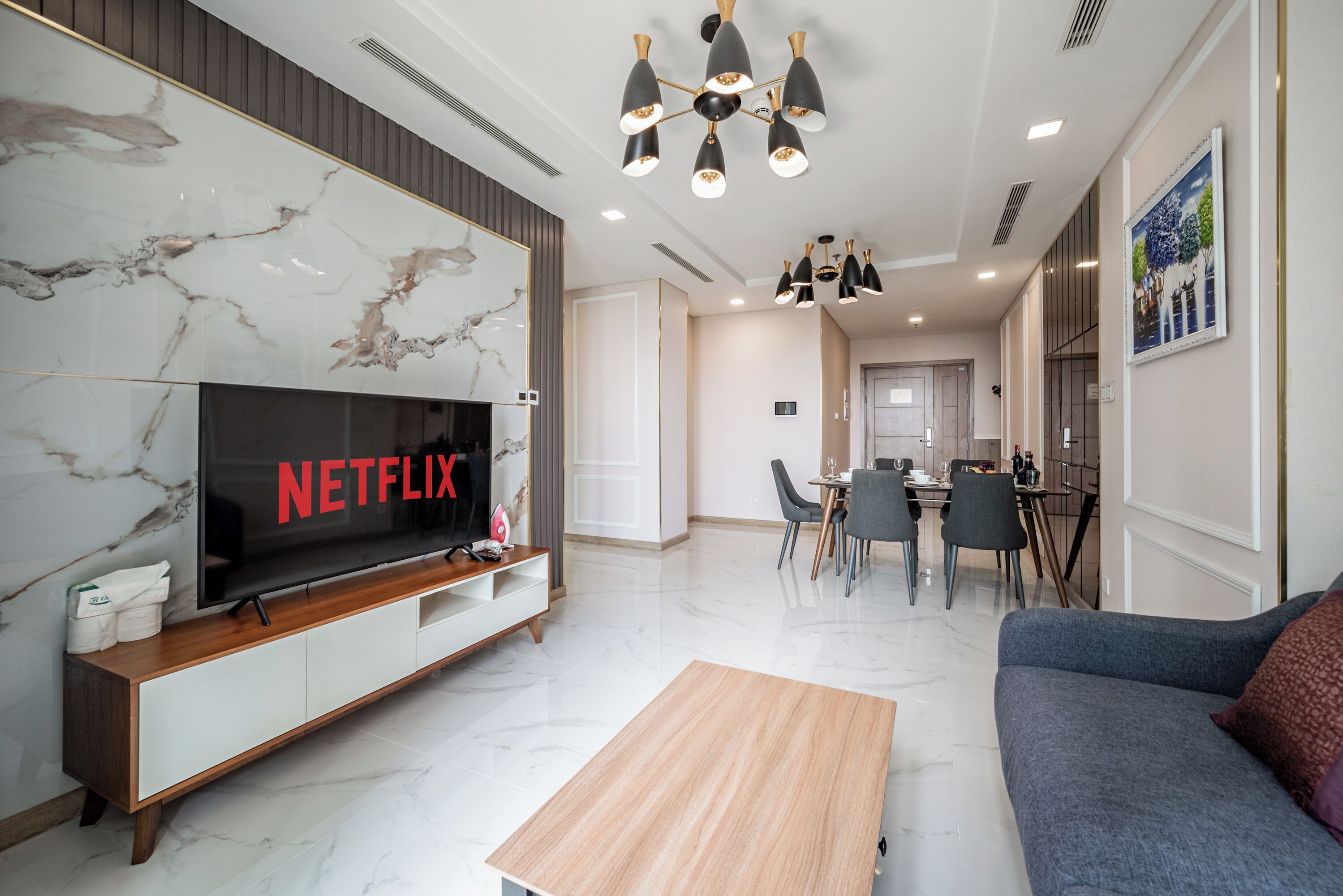 A modern living room with a large flat-screen TV, a wooden coffee table, a grey sofa, and a dining area with chairs and a table.