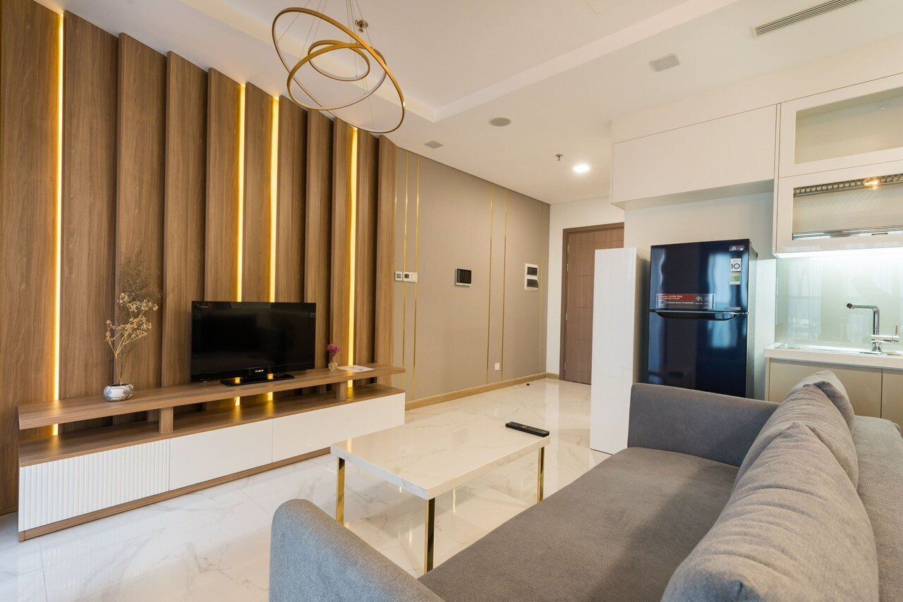 A modern living room with a flat-screen TV, a sofa, a coffee table, and a kitchen area with a refrigerator.