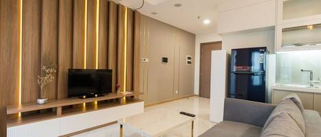A modern living room with a flat-screen TV, a sofa, a coffee table, and a kitchen area with a refrigerator.