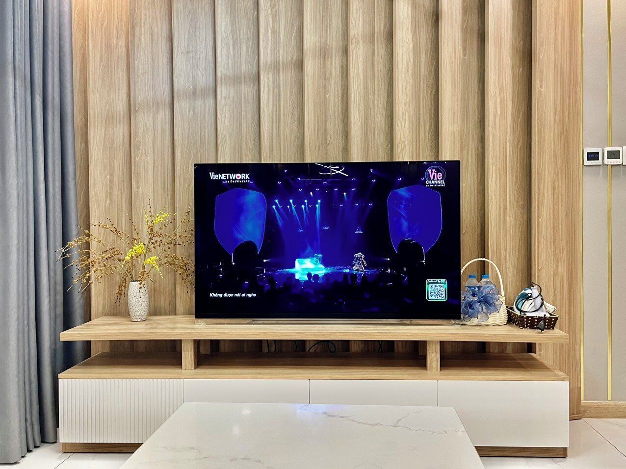 A flat-screen TV on a wooden stand with a vase of dried flowers and a basket of items.