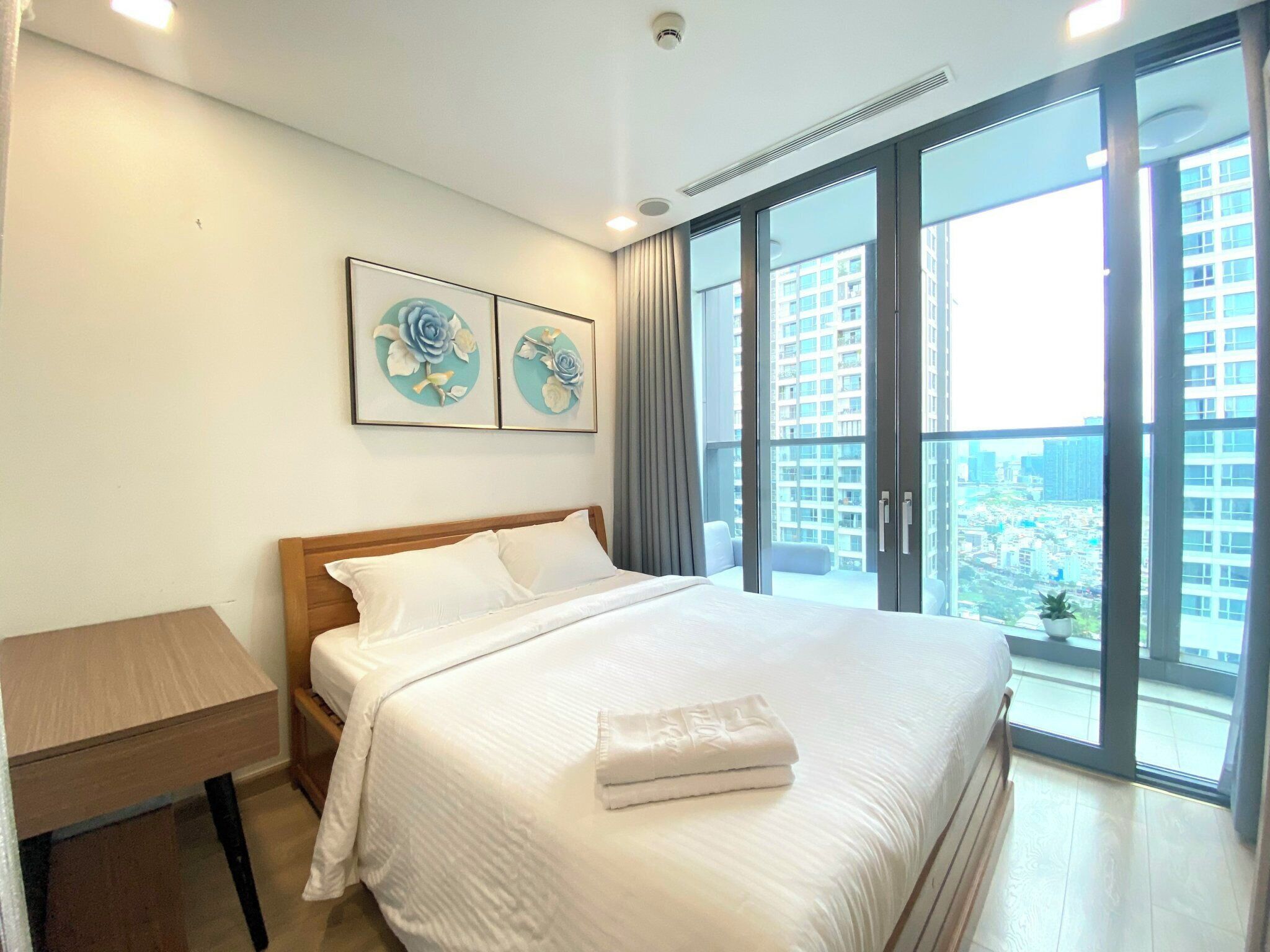 Habitación de hotel con una cama grande, una mesita de noche de madera, un escritorio y un ventanal con vistas a la ciudad.