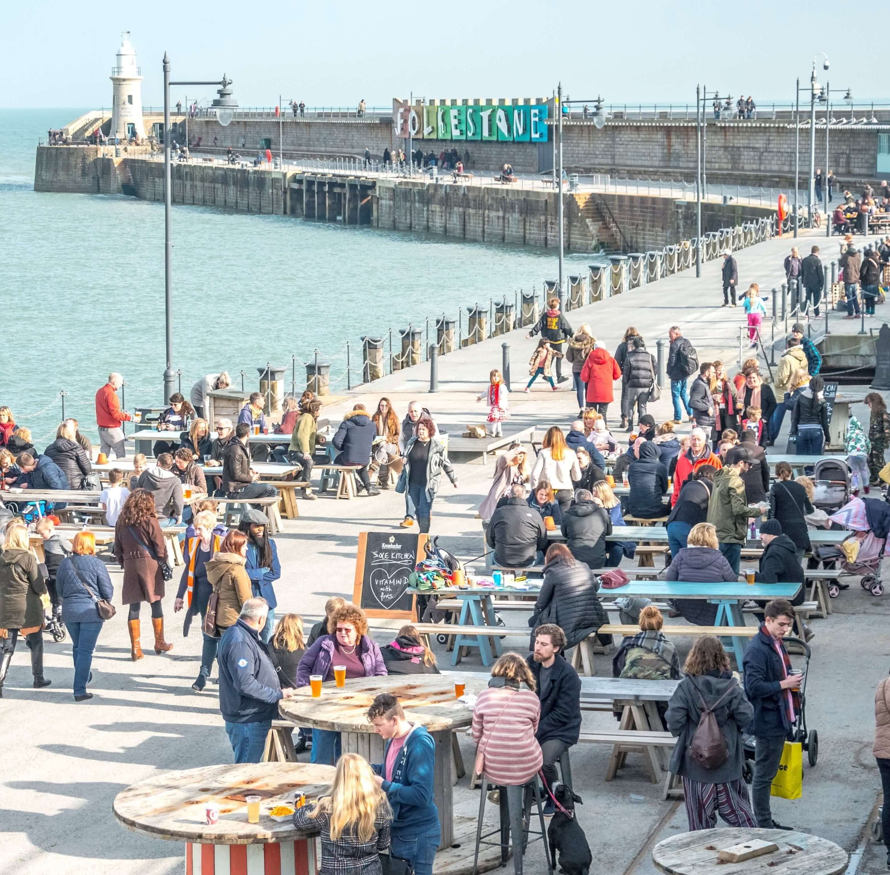 A bustling waterfront promenade with people dining at outdoor tables, a lighthouse in the background, and a sign reading "FORESTONE".