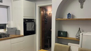 A compact kitchen with a stove, oven, and a bread container. There's a dining table with chairs and a bowl of fruit. A First Aid kit is on a shelf, and a small house decoration is on another shelf. - 1-Bedroom Apartment Sleeps 4, Parking (Hythe)