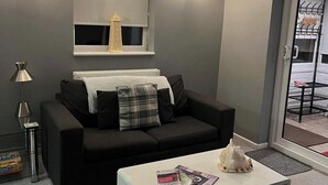 A modern living room with a black sofa, a white coffee table, and a small shelf with a decorative item. - 1-Bedroom Apartment Sleeps 4, Parking (Hythe)