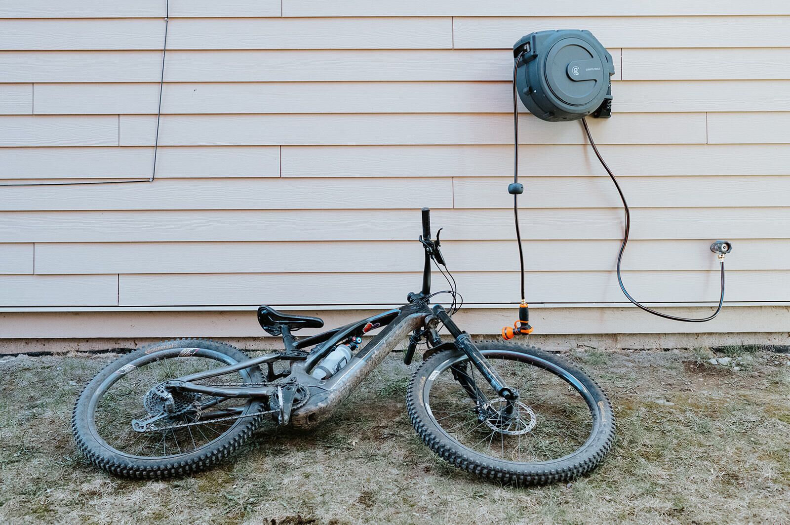 Un vélo tout-terrain est garé contre une maison, près d'une prise électrique fixée au mur et d'un tuyau d'arrosage.