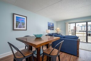A dining area with a wooden table and chairs, a blue sofa, and two framed artworks on the wall. - Ocean City Bayside Condo, Newly Renovated (Ocean City)