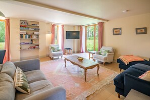 A living room with a sofa, armchairs, a coffee table, a bookshelf, a flat-screen TV, and large windows with curtains.
