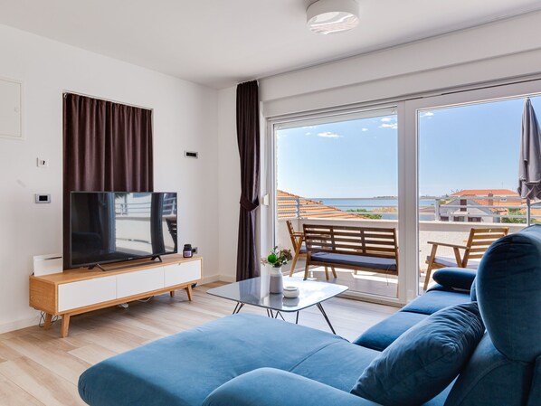 Ein modernes Wohnzimmer mit Flachbildfernseher, einer Couch, einem Couchtisch und einem Balkon mit Blick auf Meer und Gebäude.