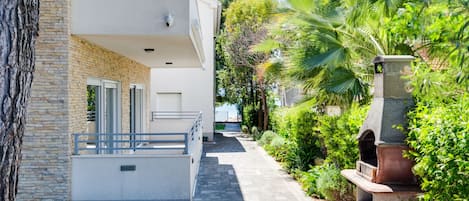 En moderne bygning med en stenfassade, en balkon med gelænder og en belagt sti, der fører til en have med palmer og en stensøjle.