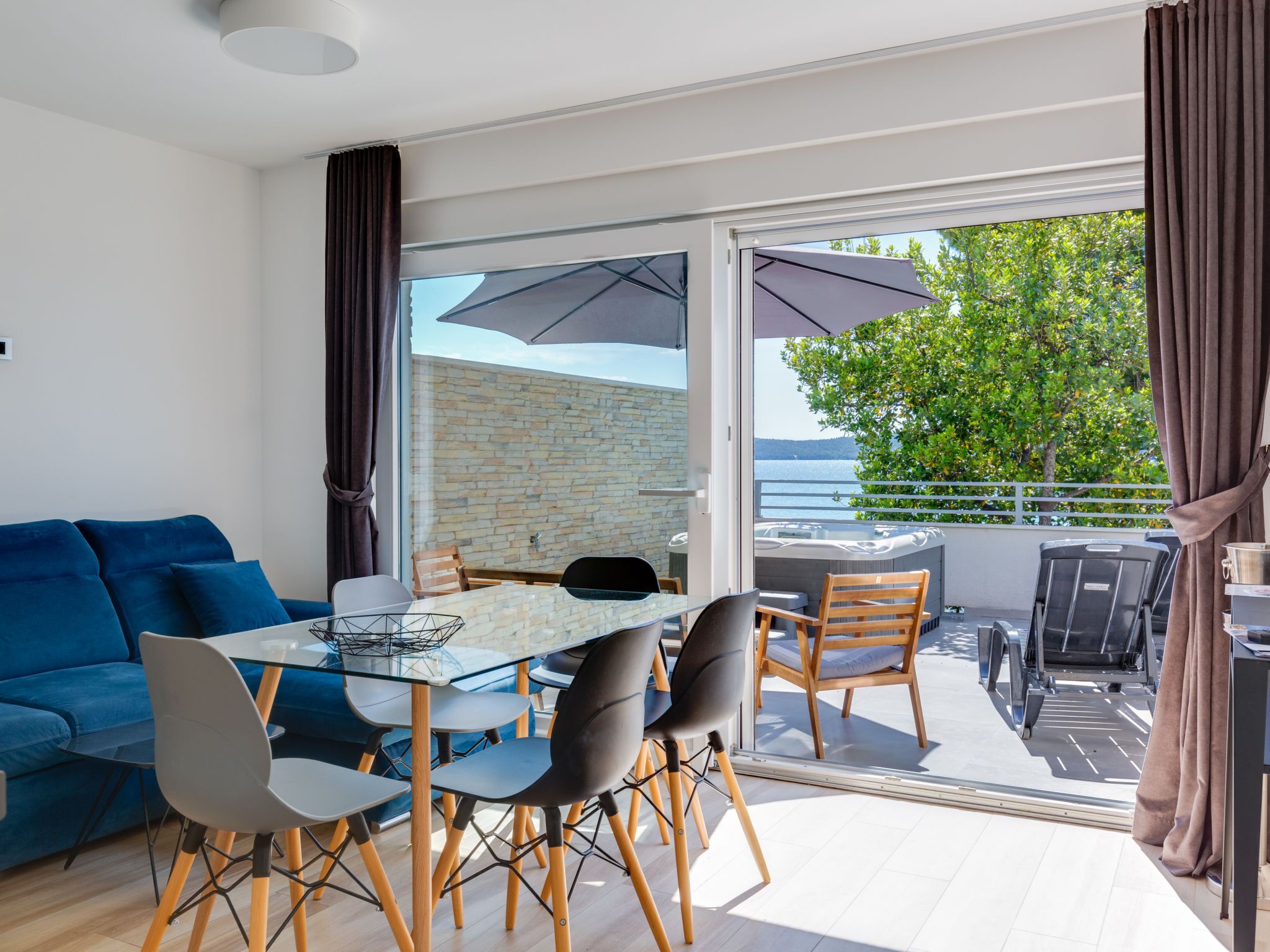 A modern living room with a glass dining table, blue sofa, and a balcony with outdoor furniture.