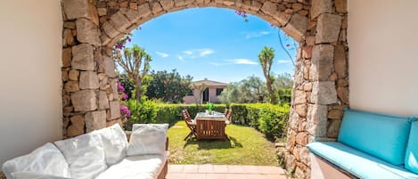 Un patio con un arco de piedra, un sofá de mimbre, un banco con cojines azules y una mesa con sillas.