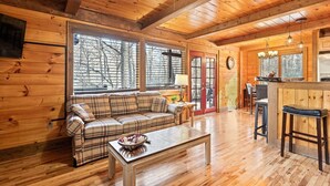 A cozy cabin interior with a wooden floor, a plaid sofa, a dining table, and a kitchen area with a bar. - Woodfern Retreat-Pet Friendly EasyCentral Location (Maggie Valley)