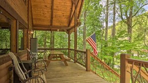A wooden deck with a picnic table and chairs, an American flag, and a view of a forested area. - Woodfern Retreat-Pet Friendly EasyCentral Location (Maggie Valley)