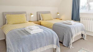 Ein Schlafzimmer mit zwei Betten, jeweils mit kariertem Bettbezug und passender Kopfstütze. Nachttischlampen, ein Fenster mit Vorhängen und ein Heizkörper sind ebenfalls vorhanden.