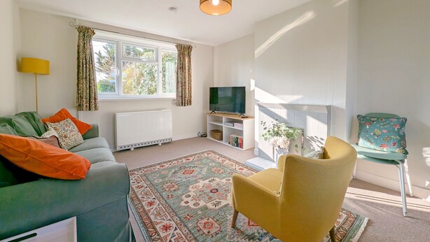 Ein Wohnzimmer mit Sofa, Sessel, Fernseher, Kamin und einem großen Fenster mit Vorhängen.