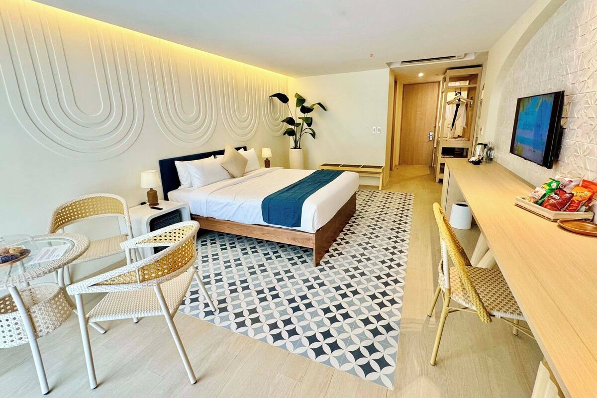 A modern hotel room with a bed, bedside table, lamp, chair, desk, and a TV.