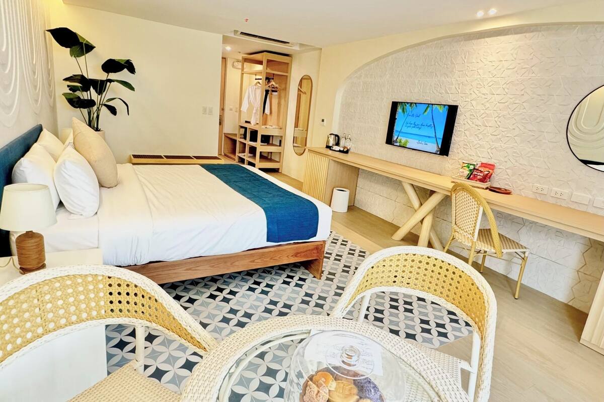 A hotel room with a bed, a desk, a TV, wicker chairs, and a small table with snacks.