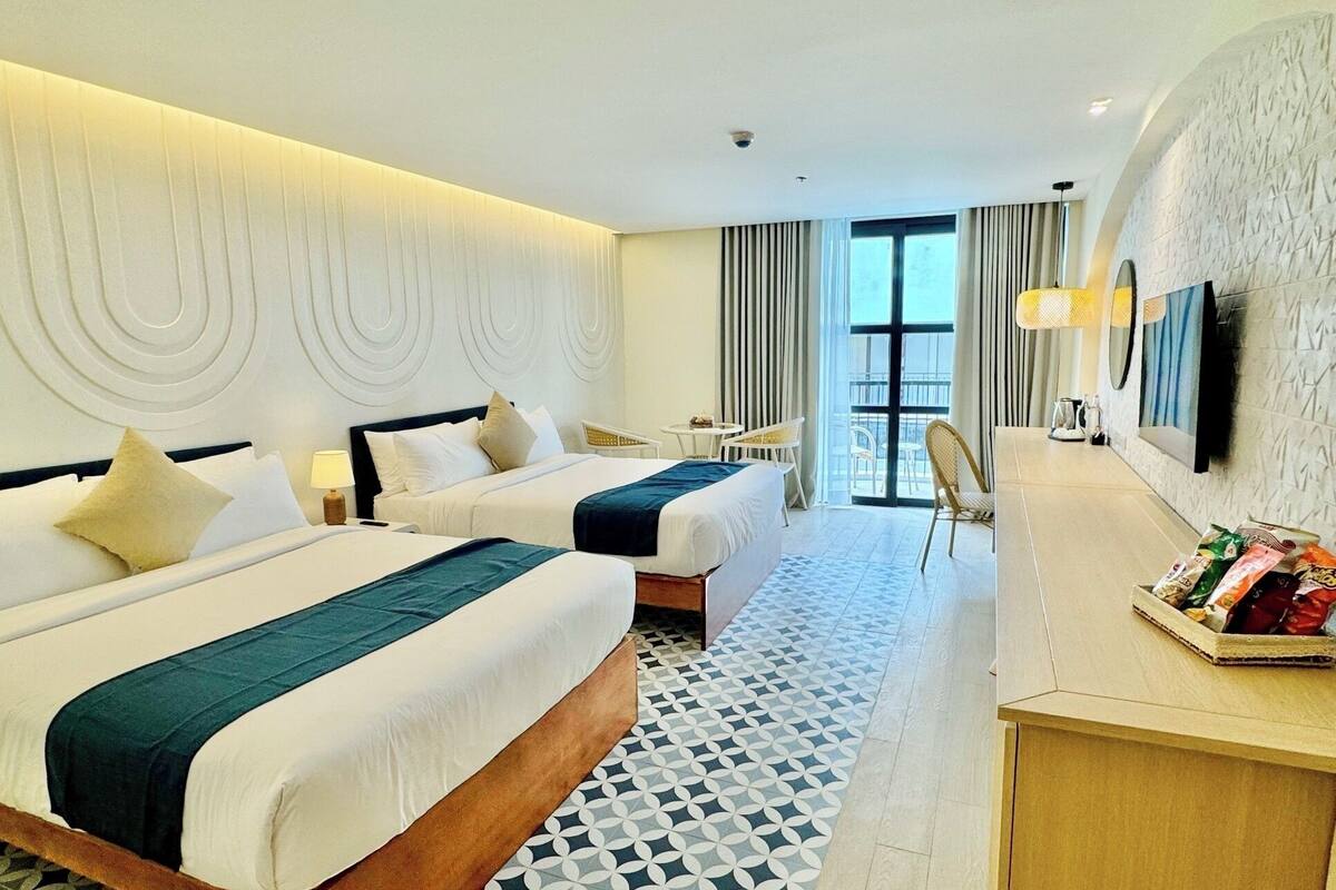 A hotel room with two beds, a desk with snacks, a TV, and a balcony with curtains.