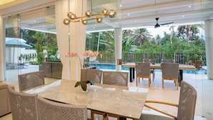 Un comedor moderno con vista a una piscina protegida por cristal, muebles de mimbre y una mesa central con un jarrón de flores.