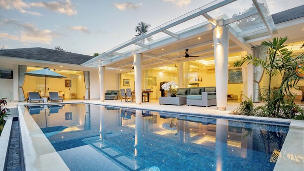 Una piscina interior moderna con agua azul cristalina, rodeada por un porche cubierto con asientos y una zona de comedor.