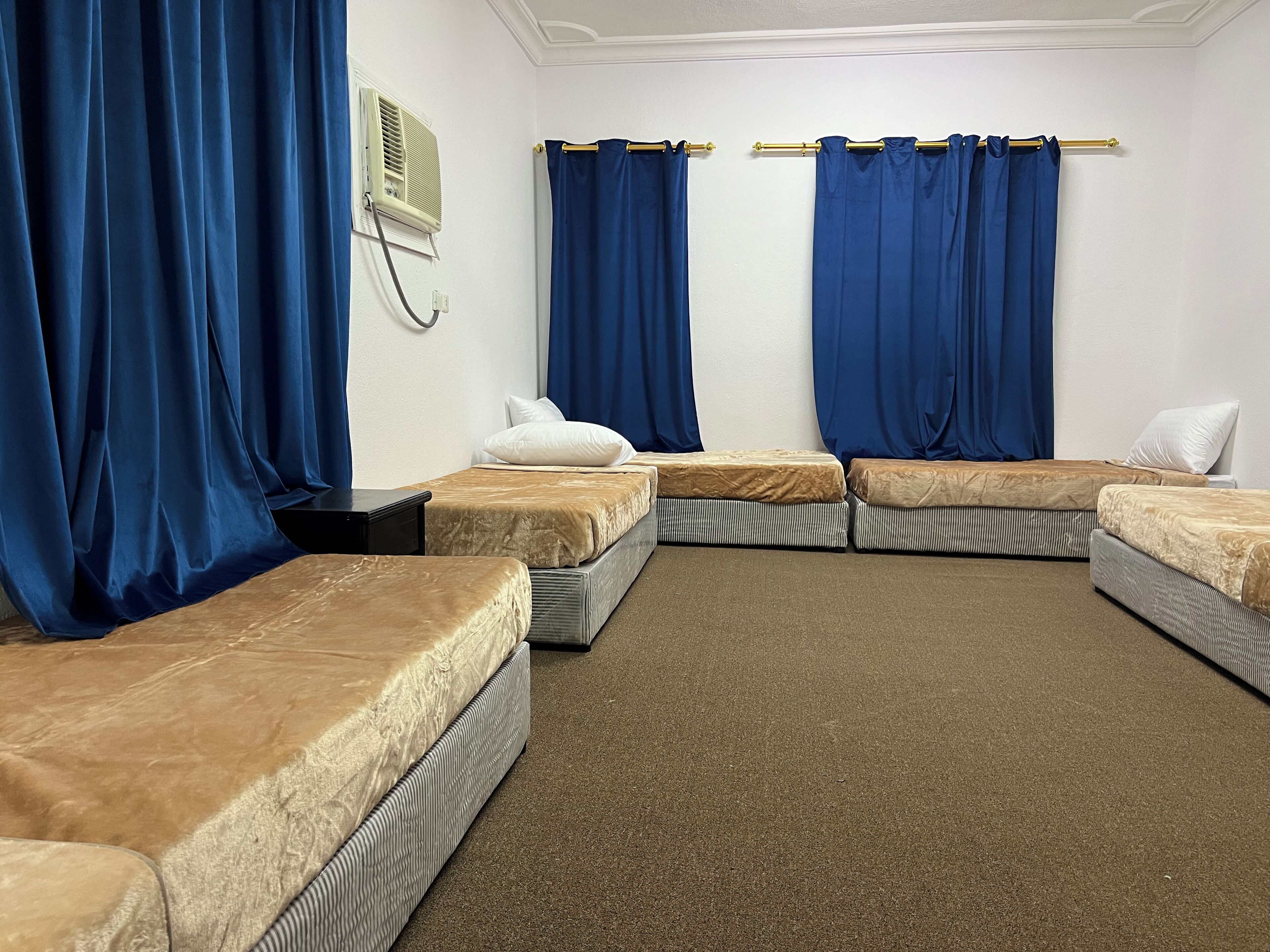 A room with four single beds, blue curtains, a wall-mounted air conditioner, and a carpeted floor.