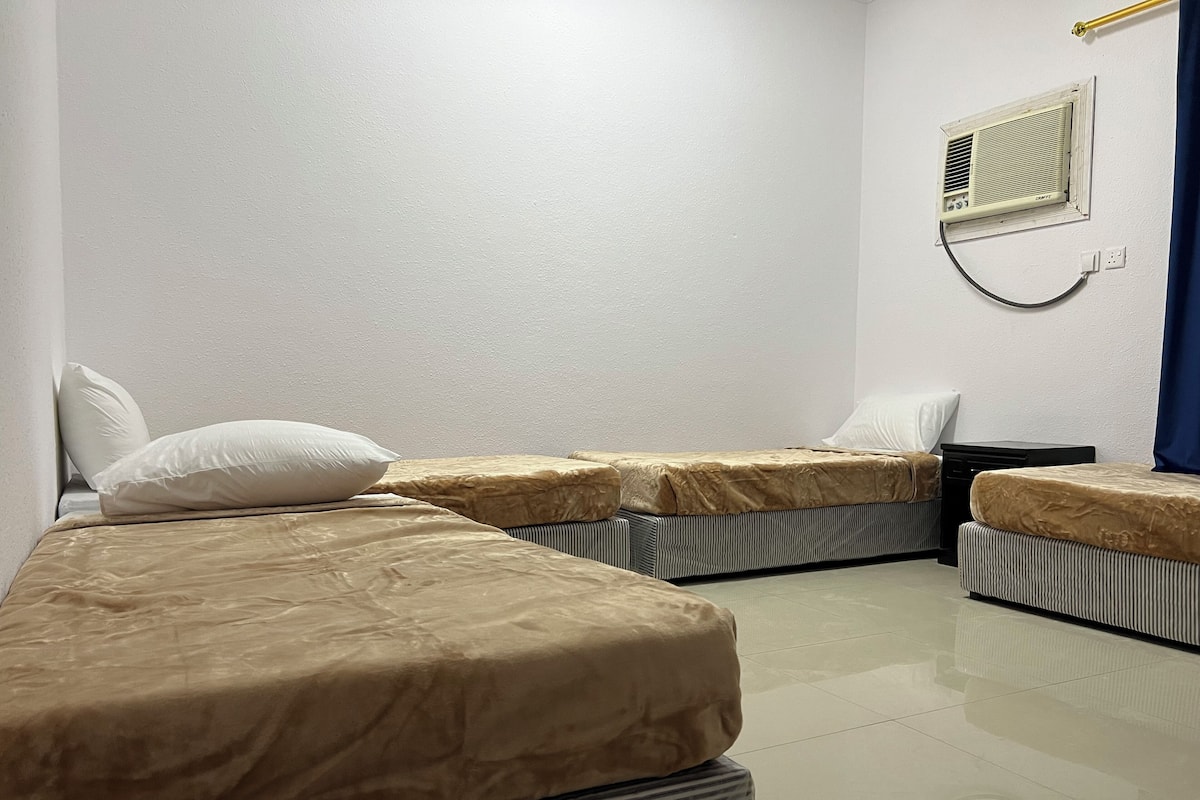 A room with two beds, each with a pillow and a blanket. There is a small table and a wall-mounted heater.