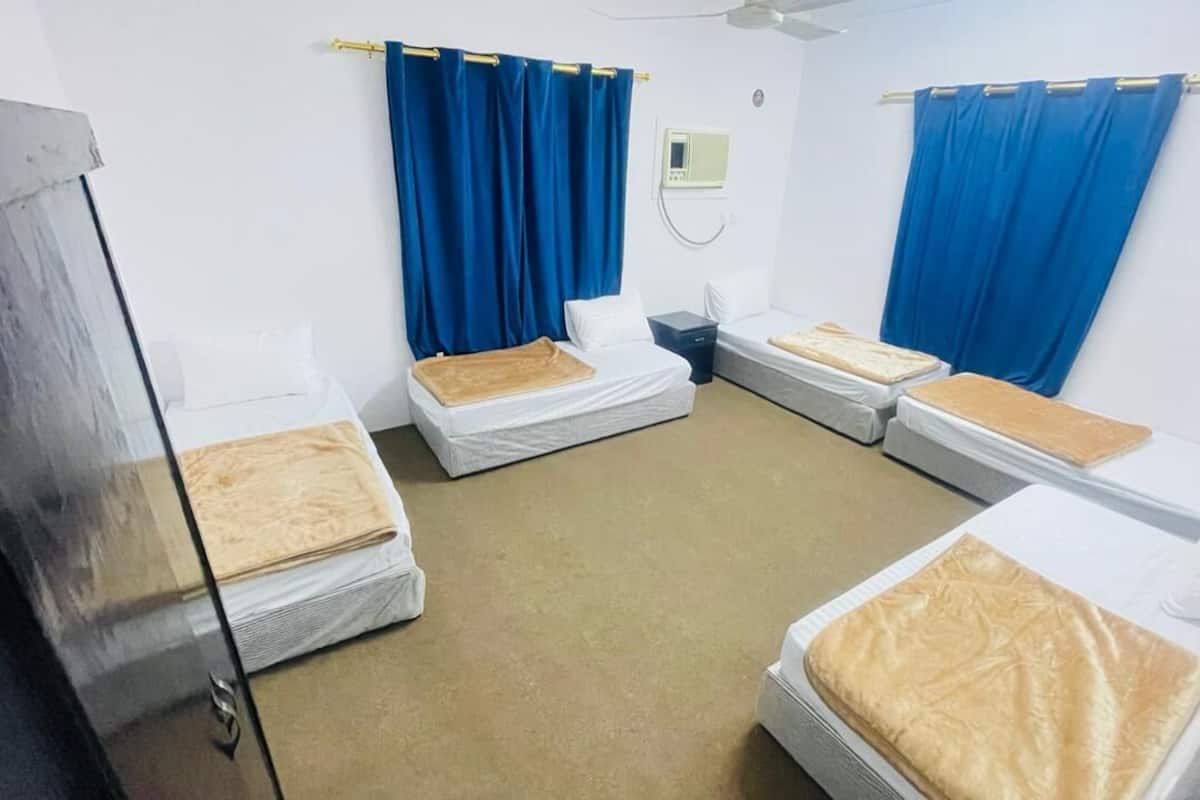 A room with four single beds, each with a white mattress and a brown blanket. Blue curtains are on the windows, and there is a ceiling fan.
