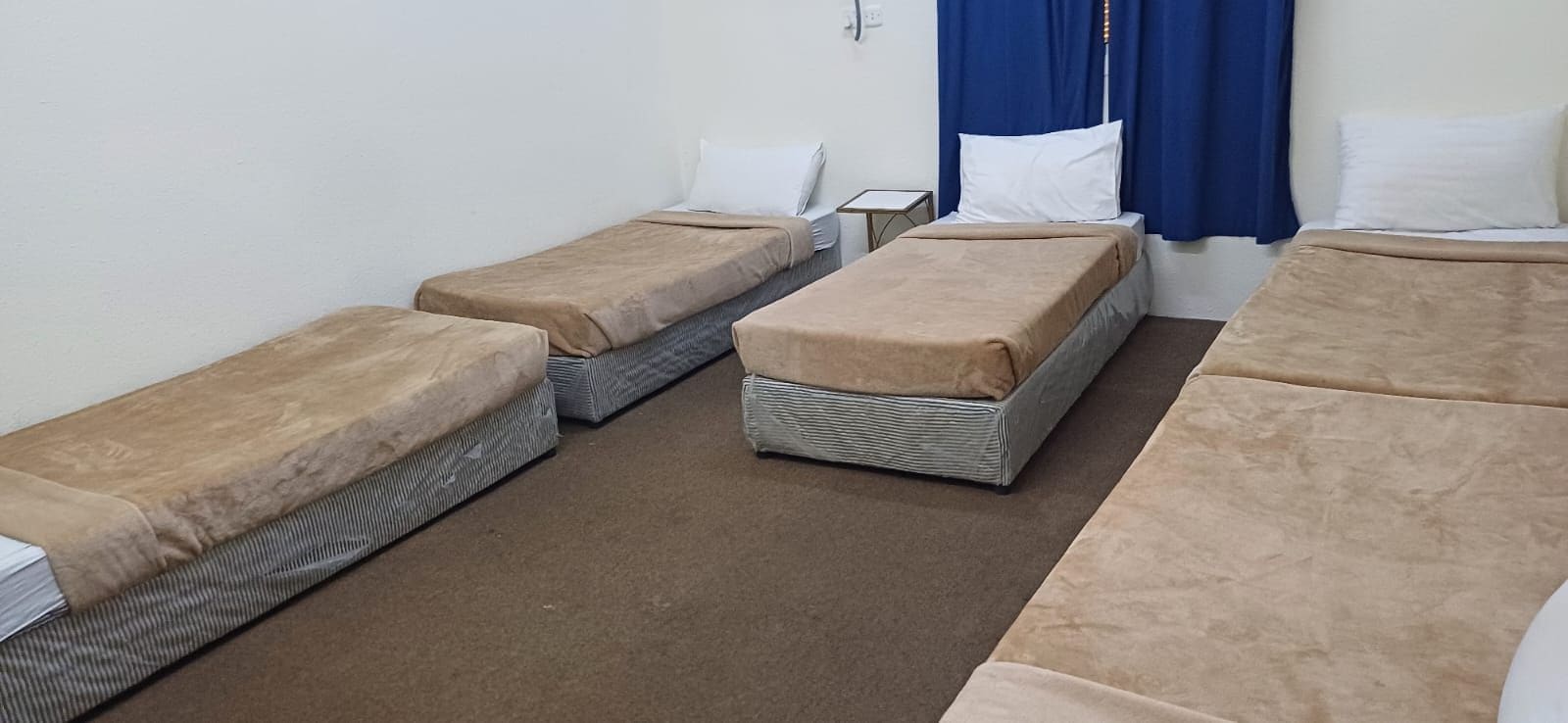 A room with four single beds arranged in a row, each with a brown mattress and white pillows. There is a small table between the beds, and a blue curtain is partially visible on the right side.
