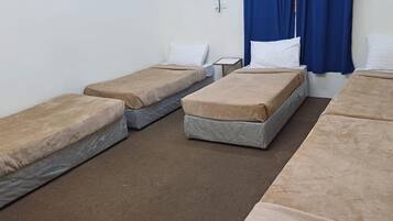 Una habitación con cuatro camas individuales dispuestas en fila, cada una con colchón marrón y almohadas blancas. Hay una mesita entre las camas, y se ve parcialmente una cortina azul al lado derecho.