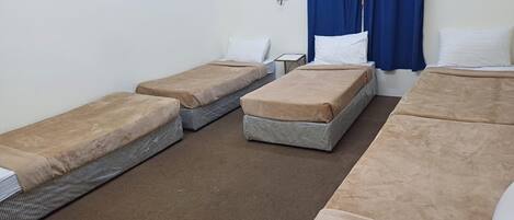 A room with four single beds arranged in a row, each with a brown mattress and white pillows. There is a small table between the beds, and a blue curtain is partially visible on the right side.