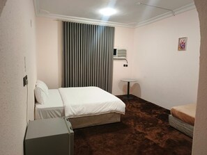 A hotel room with a bed, a small table, a bedside table, a wall-mounted air conditioner, and a window with curtains. - Jawar Al Rahma (Madinah)