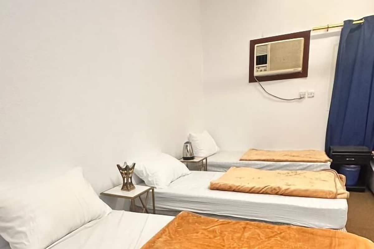 A hotel room with three beds, each with white bedding and a brown throw blanket. There is a small table with a lamp and a decorative item on it. A television is mounted on the wall above the beds.