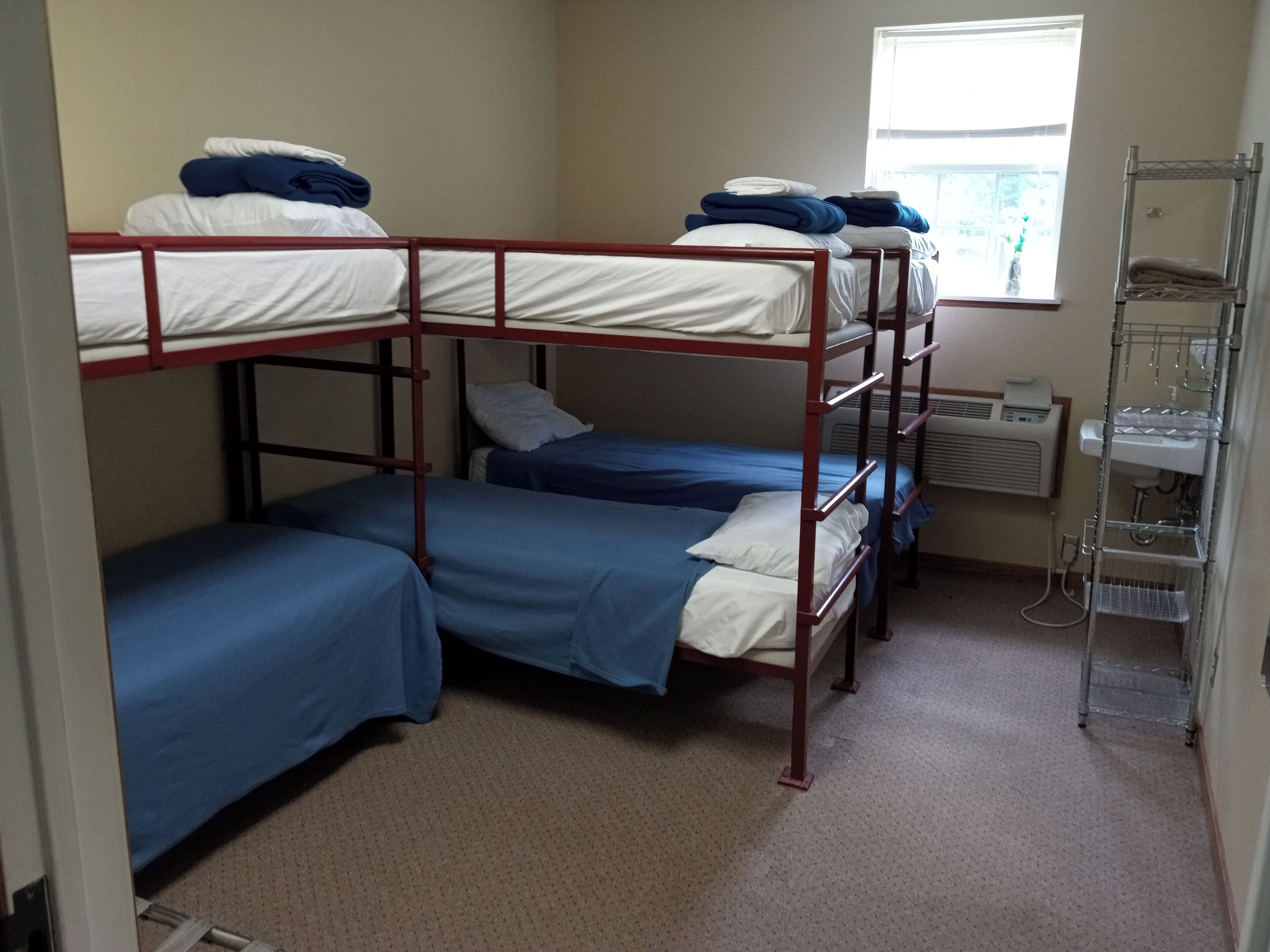 A dormitory room with bunk beds, a window with blinds, a radiator, and a metal shelf unit.