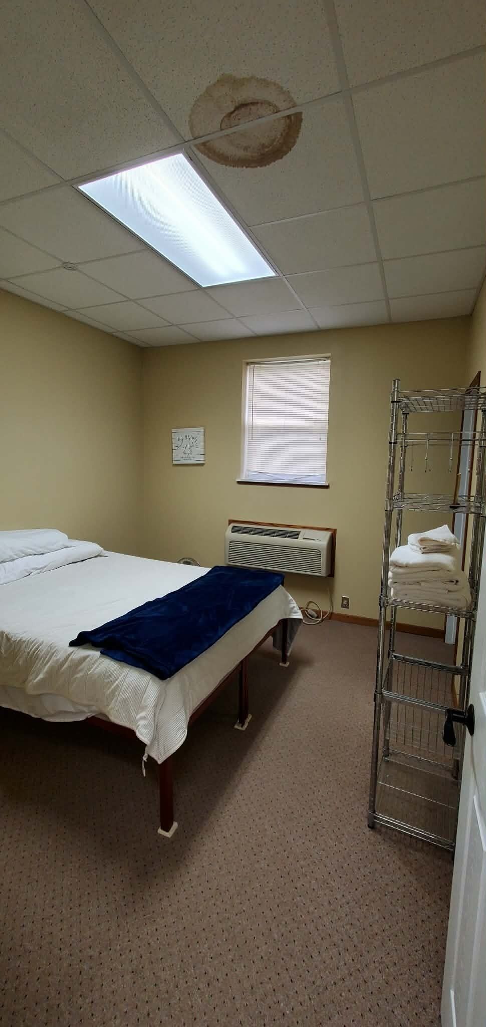 A room with a bed, a window with blinds, a radiator, and a metal shelf unit.