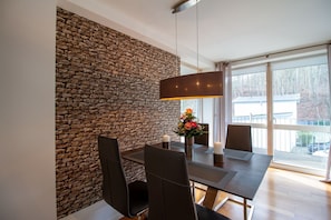 Ein stilvoller Essbereich mit Steinwand, Tisch mit Stühlen, einer Vase mit Blumen und großen Fenstern mit Blick ins Freie.