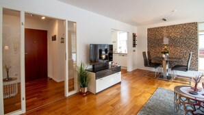 A modern living room with a flat-screen TV, a dining area with a stone accent wall, and a hallway leading to another room. - Ocean Club - Exklusive Ferienwohnung: mit Balkon, Direktem Ostseeblick & Strandzugang (Timmendorfer Strand)