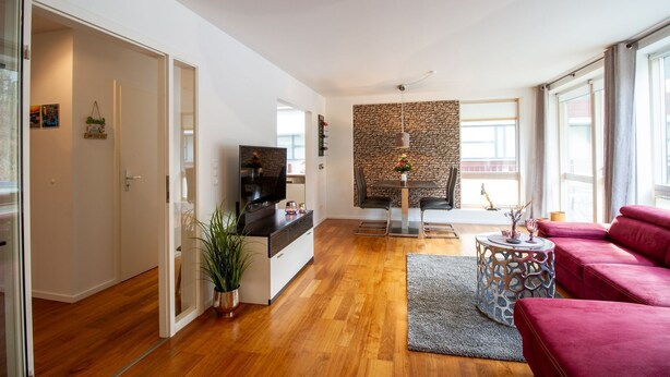 A spacious living room with a flat-screen TV, a dining area with a stone accent wall, and a red sofa. - Ocean Club - Exklusive Ferienwohnung: mit Balkon, Direktem Ostseeblick & Strandzugang (Timmendorfer Strand)
