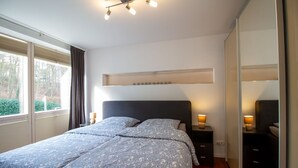 A bedroom with a large bed, bedside tables with lamps, a built-in shelf above the bed, and a view through a window. - Ocean Club - Exklusive Ferienwohnung: mit Balkon, Direktem Ostseeblick & Strandzugang (Timmendorfer Strand)