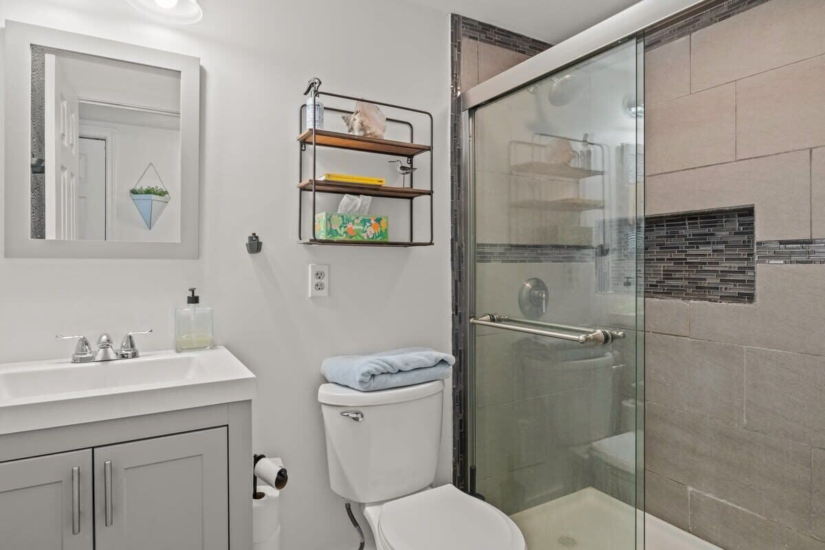 A modern bathroom with a glass shower enclosure, a white toilet, and a vanity with a mirror and shelves.