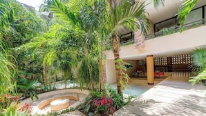 A lush indoor garden with a central water feature, surrounded by various tropical plants and a seating area with red chairs. - Stylish Playa Condo: Rooftop Pool, Gym, Near Beach (Playa del Carmen)