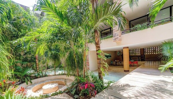 Un jardin intérieur luxuriant, avec une fontaine centrale, entouré de différentes plantes tropicales et agrémenté d’un espace détente avec des chaises rouges.