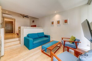 Un salon moderne avec un canapé bleu, une table basse en bois et un fauteuil bleu. On y trouve également un ventilateur de plafond, un téléviseur et des œuvres d’art accrochées au mur.