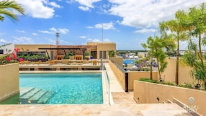 A rooftop pool area with clear blue water, a stone wall, and a lounge with chairs and tables. - Stylish Playa Condo: Rooftop Pool, Gym, Near Beach (Playa del Carmen)