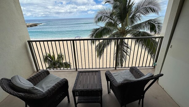 A balcony with wicker furniture, a view of a beach, and a palm tree.